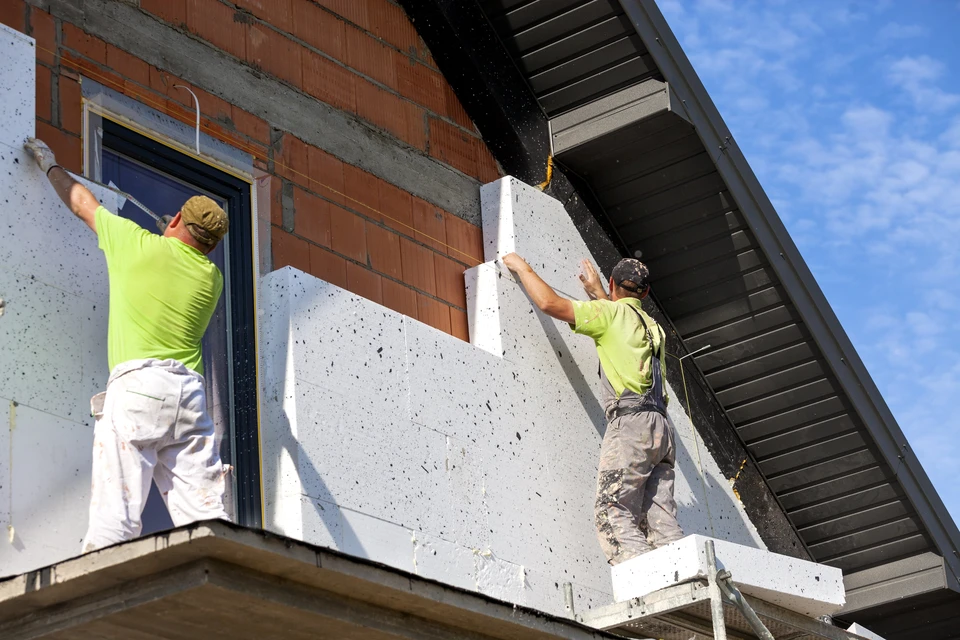 Deux ouvriers en v&ecirc;tements haute visibilit&eacute; installent des panneaux d'isolation thermique ext&eacute;rieure sur fa&ccedil;ade de maison individuelle